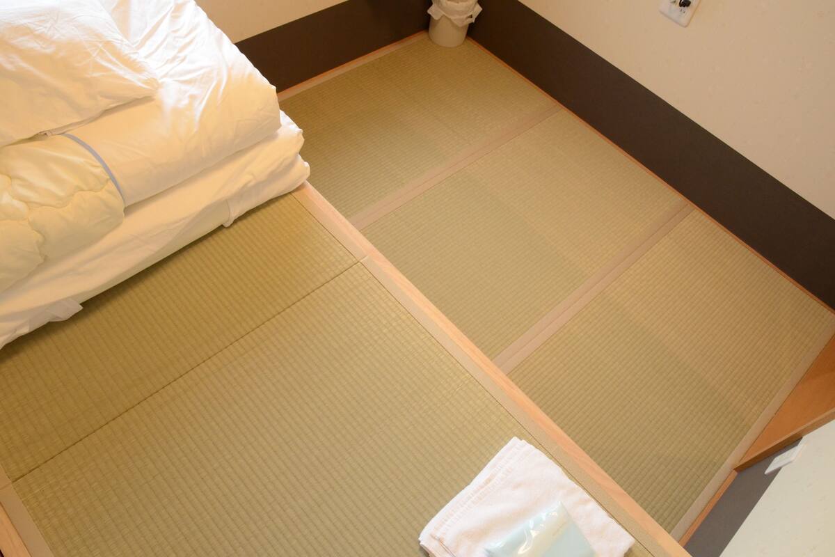 Ein Zimmer mit Tatami-Matten, einem Bett mit weißer Bettwäsche und einem kleinen weißen Mülleimer.