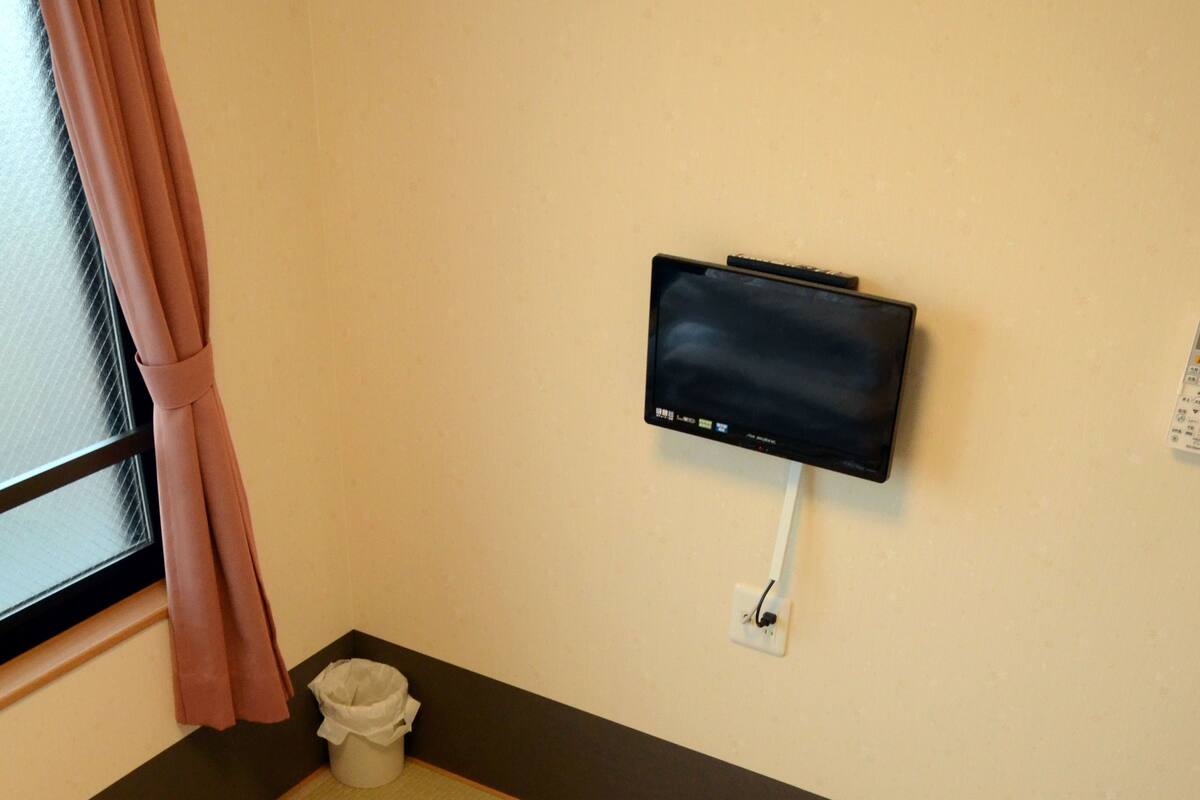 Ein Zimmer mit Tatami-Boden, einem an der Wand montierten Fernseher, einem Mülleimer und einem Fenster mit Vorhängen.