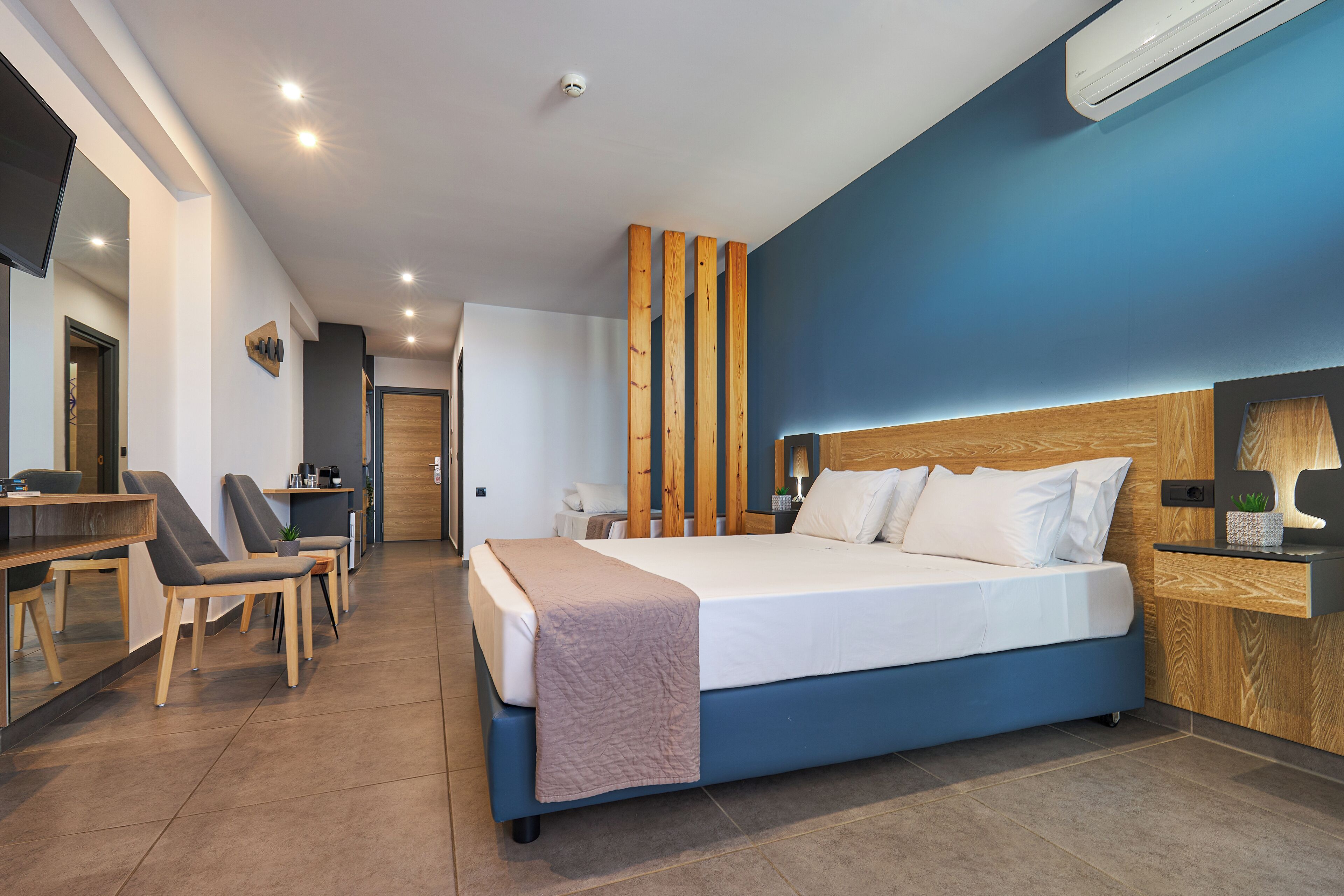Ein modernes Hotelzimmer mit einem großen Bett, einem Schreibtisch und einem Sessel. Es gibt ein Holz-Kopfteil, einen Nachttisch mit Lampe sowie eine Wand-Klimaanlage.