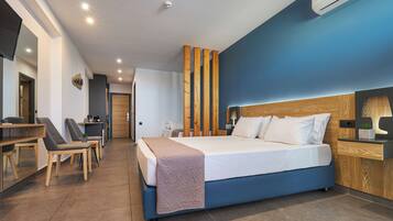 Ein modernes Hotelzimmer mit einem großen Bett, einem Schreibtisch und einem Sessel. Es gibt ein Holz-Kopfteil, einen Nachttisch mit Lampe sowie eine Wand-Klimaanlage.