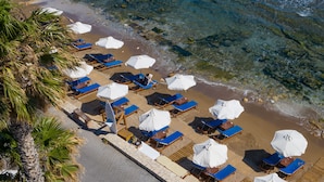 Luchtbeeld van een strand met rijen ligbedden en parasols, helderblauw water en palmbomen.