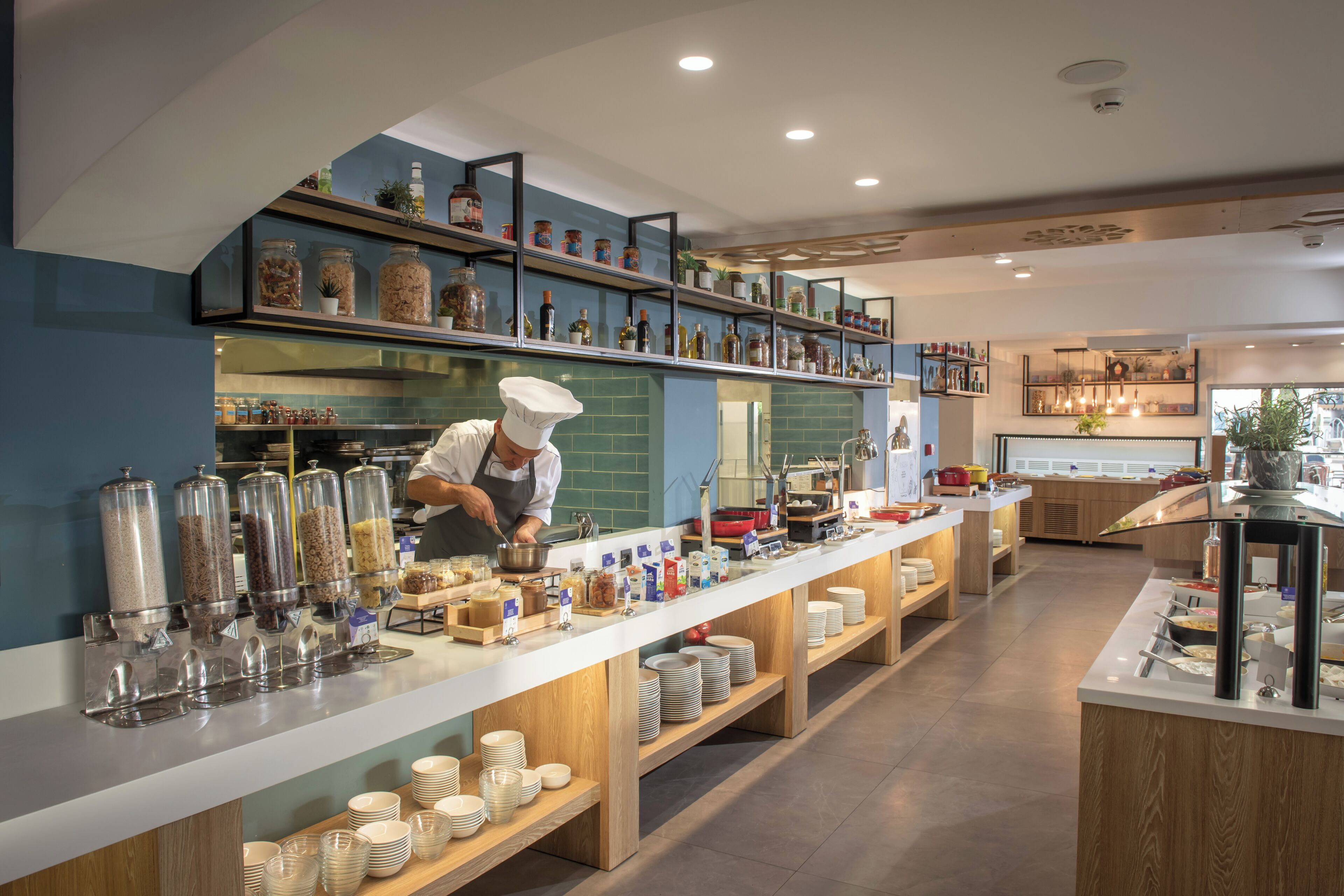 A modern kitchen with a chef preparing food, a long counter with food containers, and shelves with kitchen items.