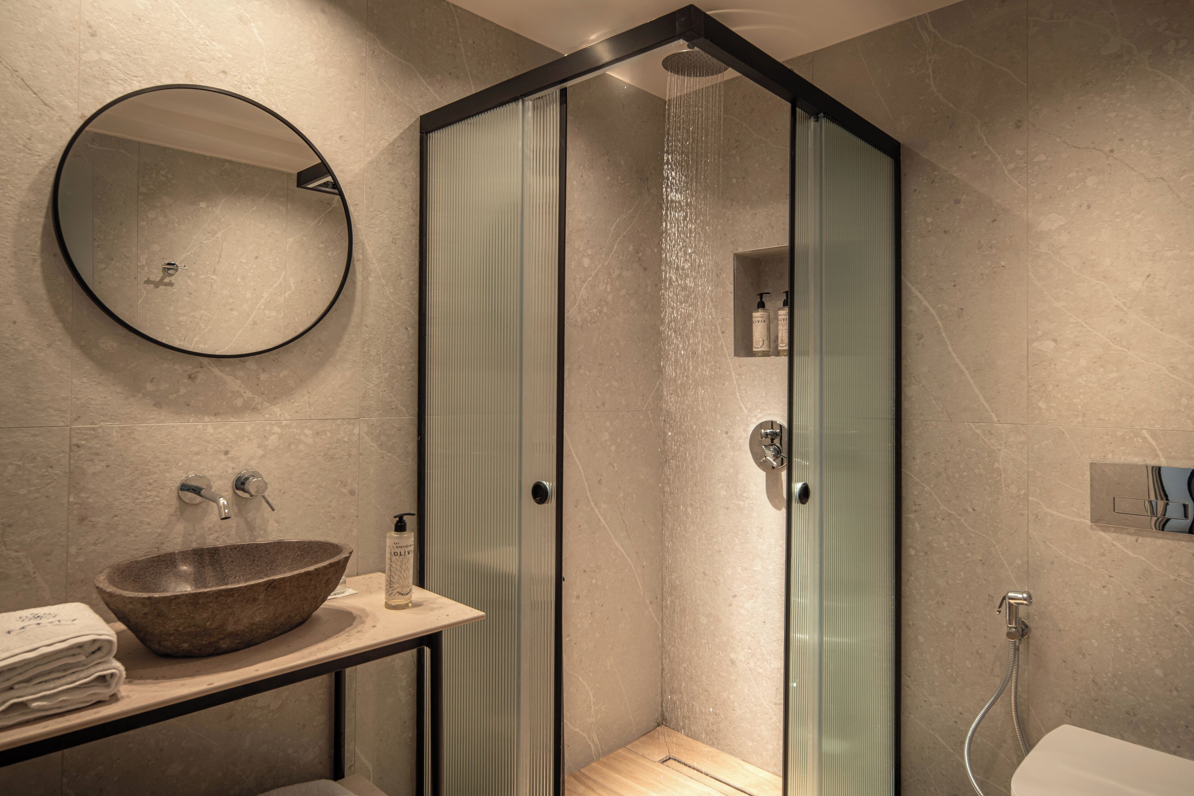 A modern bathroom with a round mirror, a wooden bowl sink, a glass-enclosed shower, and a toilet.