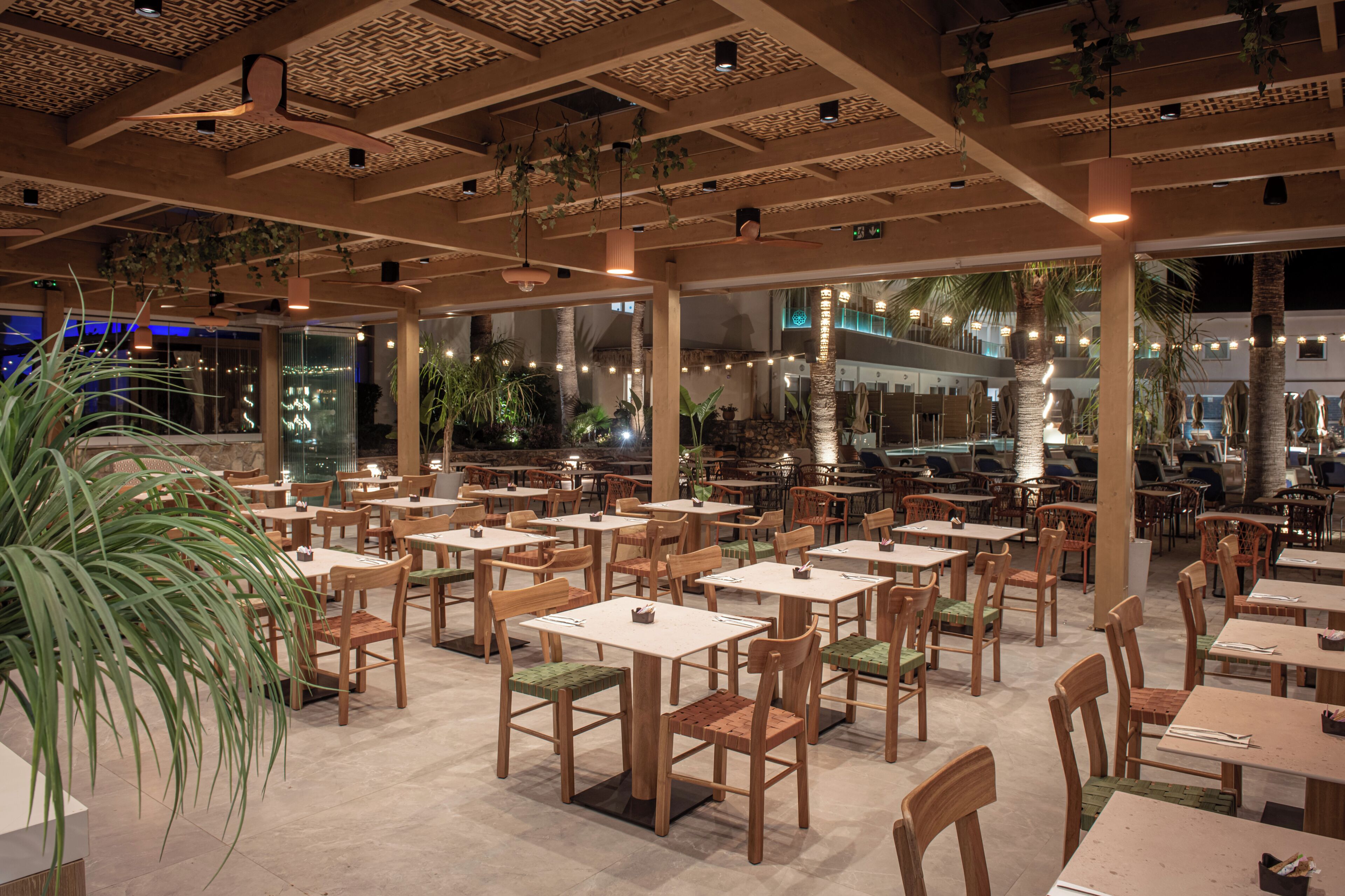 A spacious indoor dining area with multiple tables and chairs, large windows, and hanging plants.