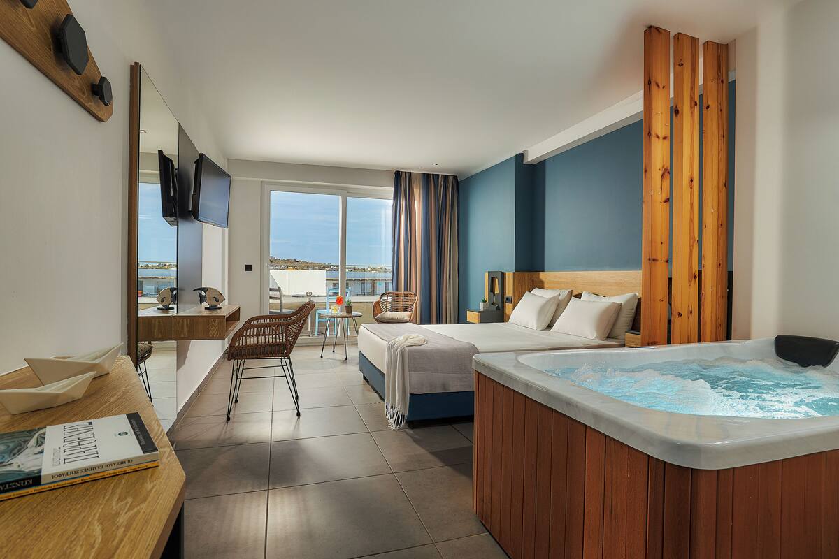 Ein modernes Hotelzimmer mit einem großen Bett, einem hölzernen Kopfteil, einer Whirlwanne, einem Schreibtisch mit Stuhl und Blick auf das Meer.