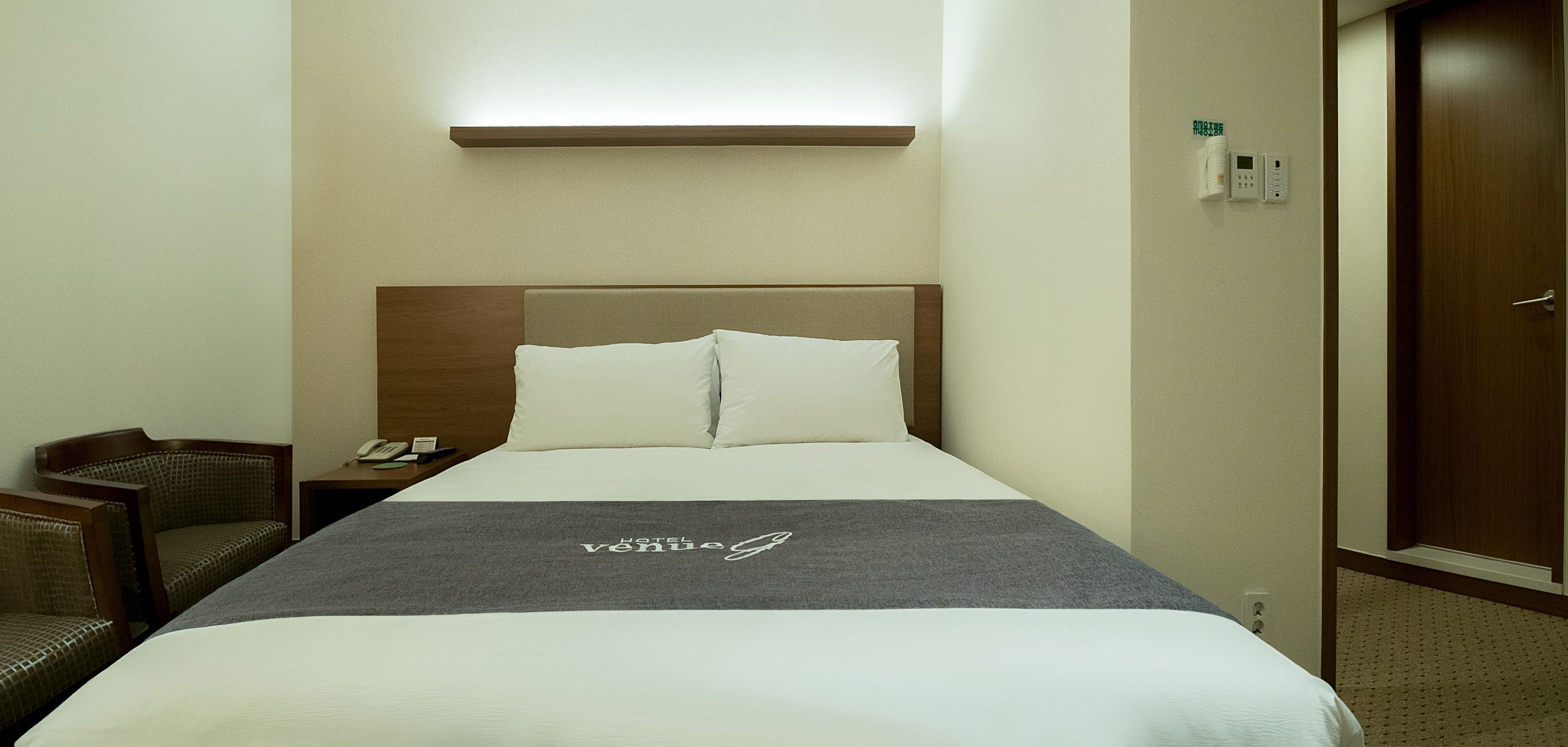 Quarto de hotel com cama, mesa de cabeceira, cadeira, telefone fixado na parede e uma porta.