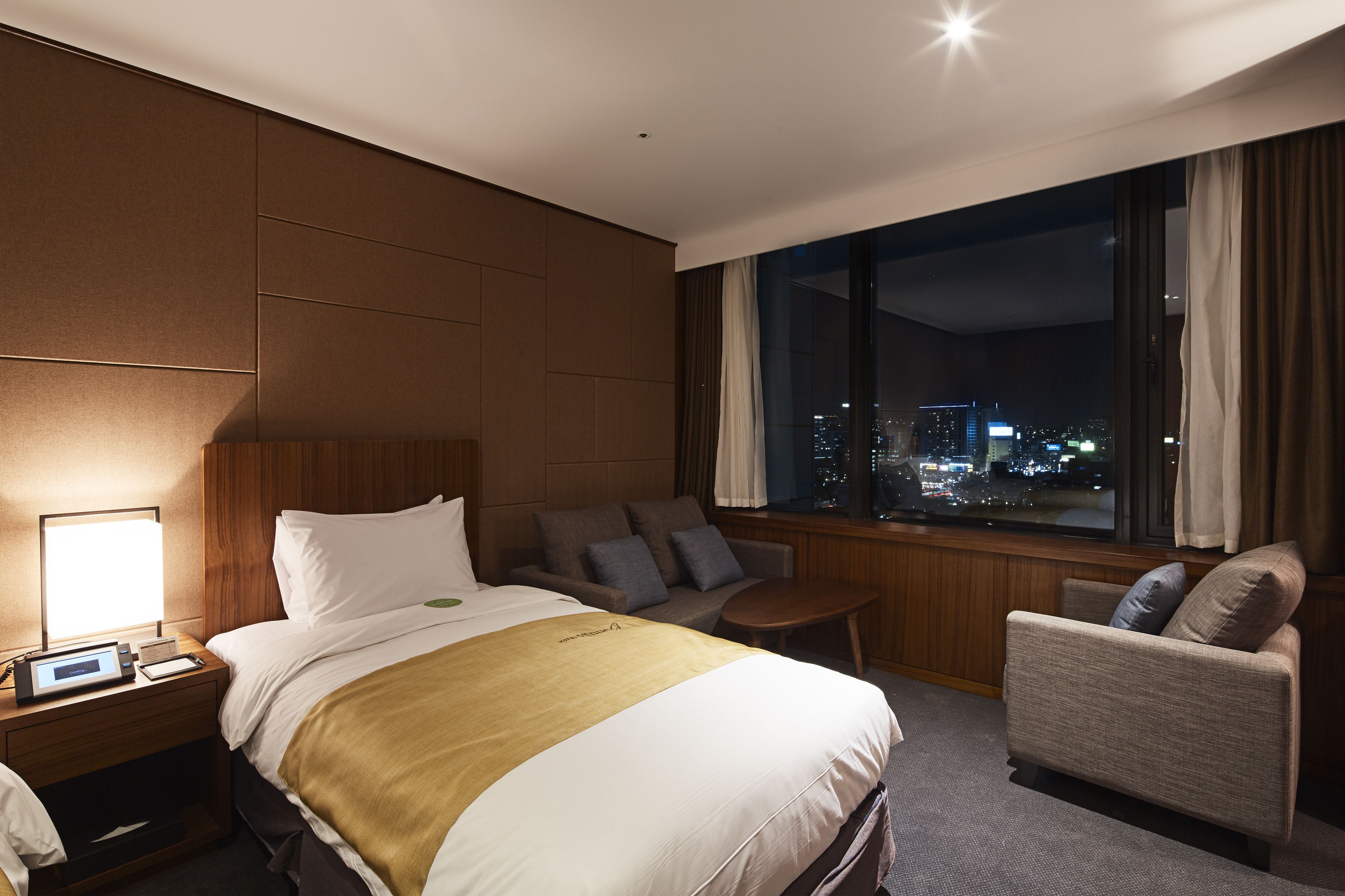 Una habitación de hotel con una cama grande, una zona de estar con sofá y sillón, una mesita y una ventana con vistas a la ciudad.