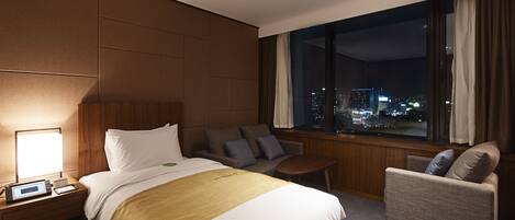 Una habitación de hotel con una cama grande, una zona de estar con sofá y sillón, una mesita y una ventana con vistas a la ciudad.