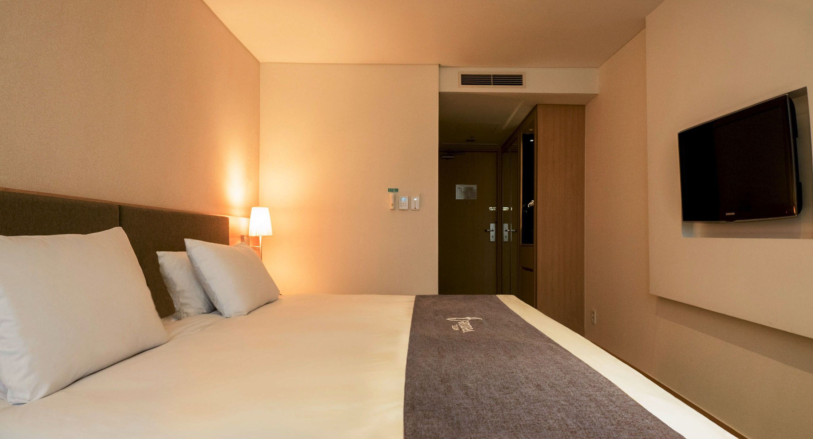 A hotel room with a bed, a TV mounted on the wall, and a lamp on the nightstand.