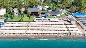 Vue aérienne d’un complexe hôtelier situé en bord de mer, avec de nombreux transats et parasols, une piscine et un restaurant.