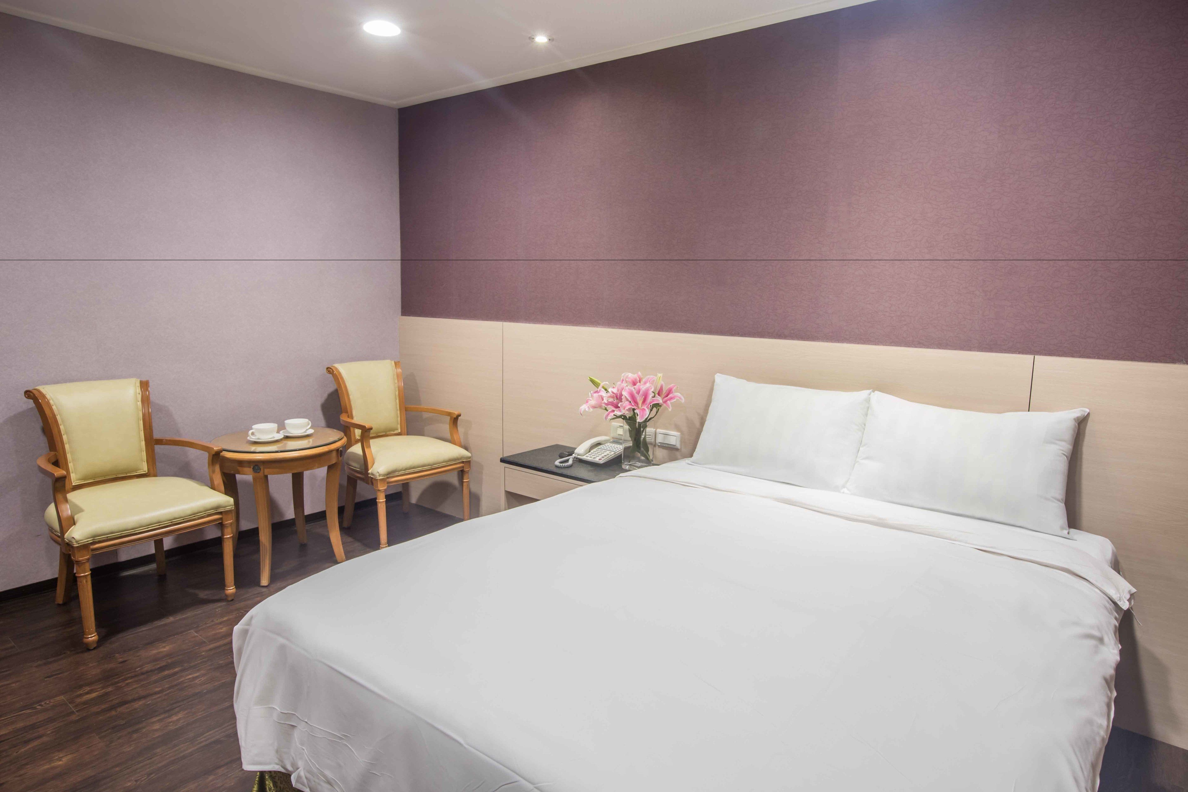 A hotel room with a large bed, two chairs, a small table, a vase with flowers, and a telephone on the nightstand.