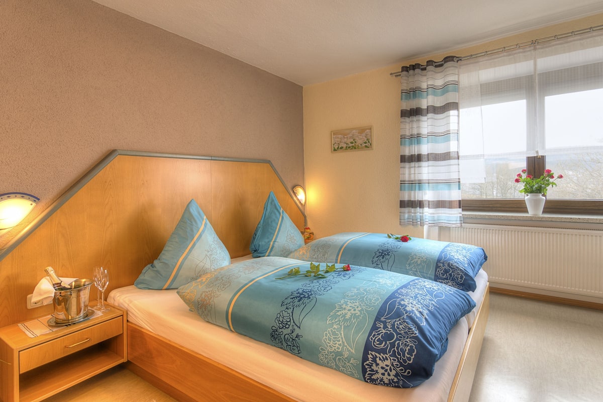 A bedroom with a wooden headboard, a bedside table with a lamp, a window with curtains, and a view of trees outside.