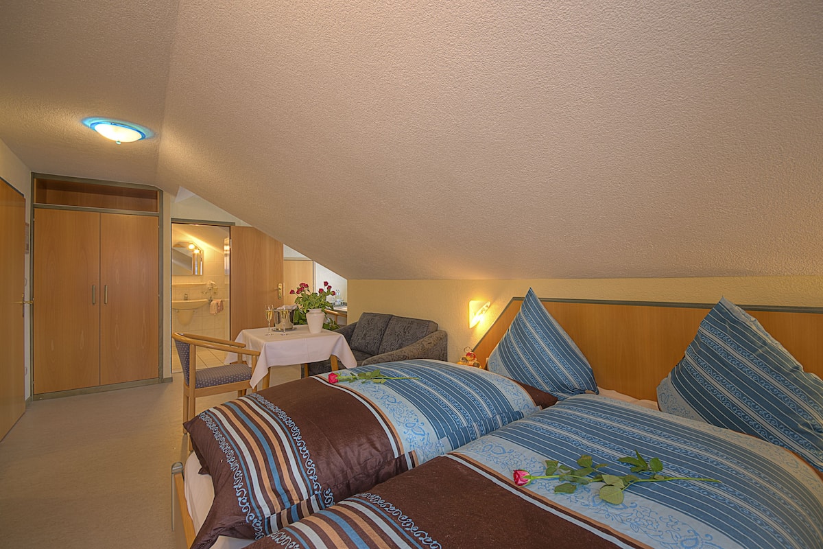 A bedroom with a bed, a chair, a table, and a bathroom in the background.
