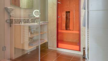 Un baño moderno con cabina de ducha de vidrio, tocador de madera y puerta enmarcada en rojo que da acceso a la zona de la ducha.
