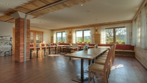 A spacious dining area with long tables and benches, large windows with curtains, and a brick column. - Landhotel Goldenes Kreuz (Moosbach)