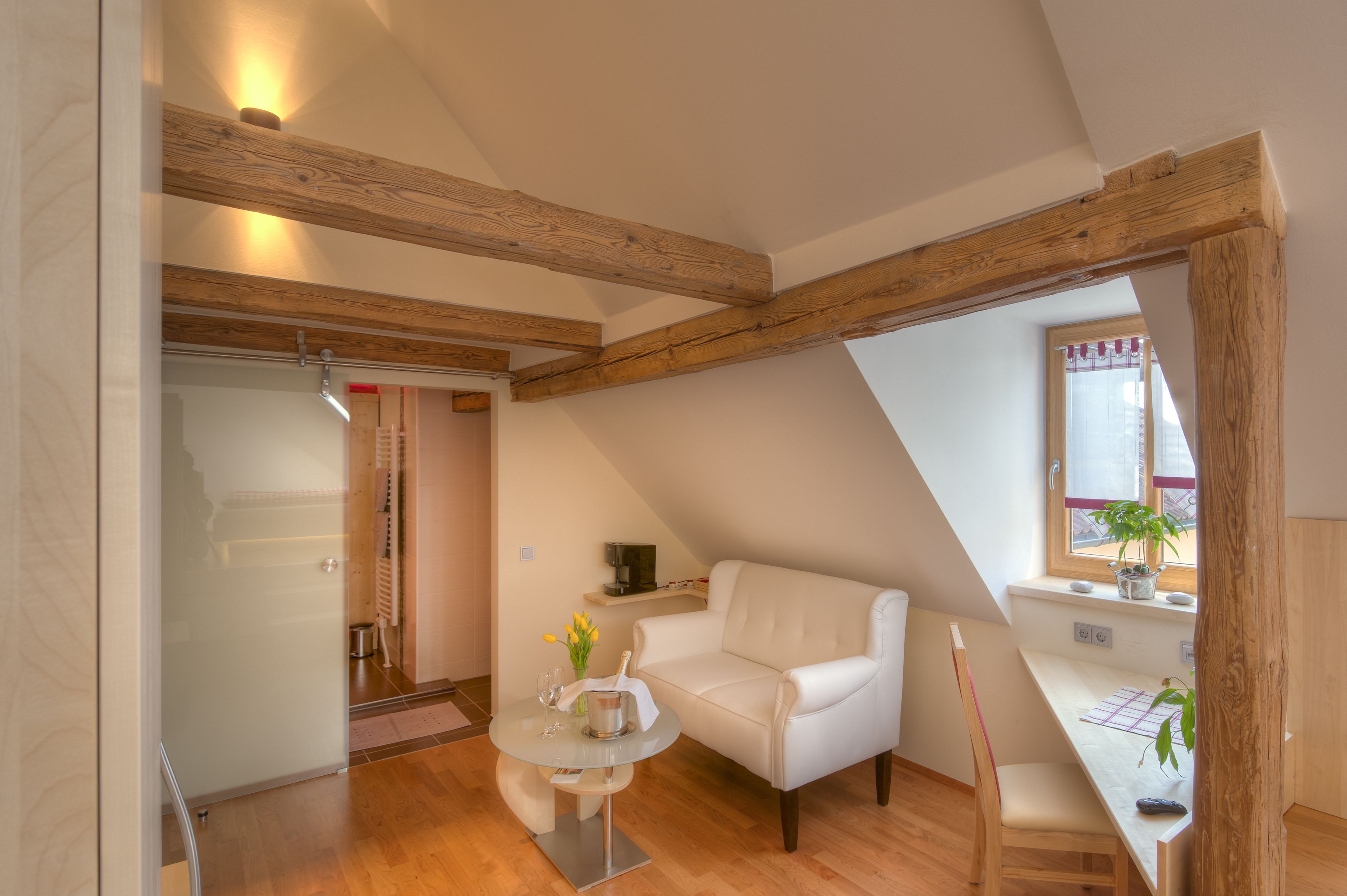 A modern loft bedroom with exposed wooden beams, a white sofa, a small round table with a vase of flowers, and a window with a view.
