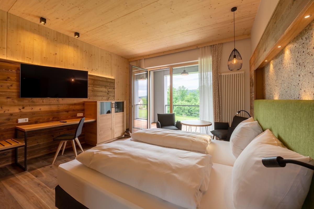 A modern hotel room with a wooden ceiling, a large flat-screen TV, a bed with white linens, a desk with a chair, and a balcony with a view of greenery.