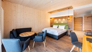 A modern hotel room with a wooden ceiling, a bed with white bedding, a sofa, two chairs, a small round table, and a wooden desk. - Landhotel Goldenes Kreuz (Moosbach)