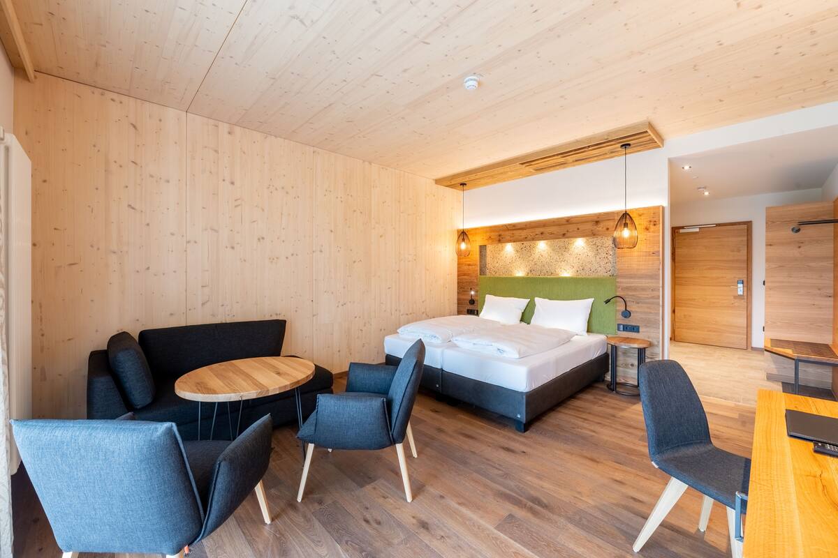 A modern hotel room with a wooden ceiling, a bed with white bedding, a sofa, two chairs, a small round table, and a wooden desk.