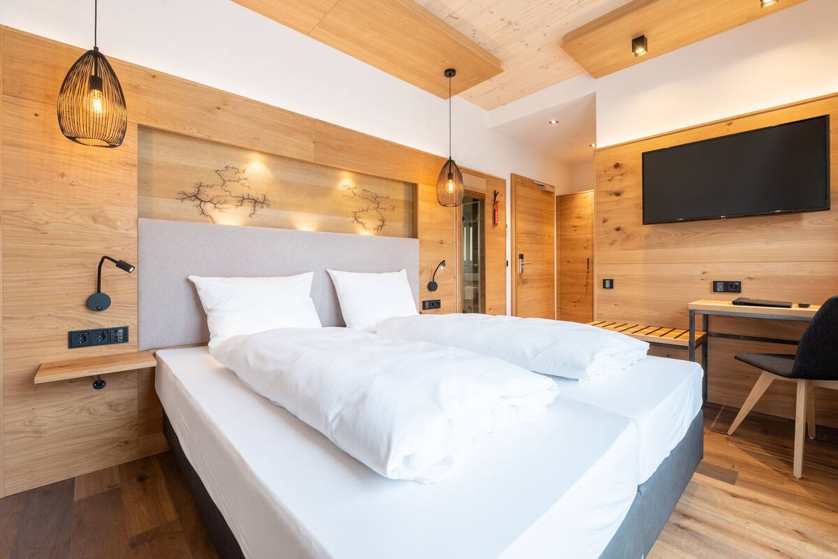 A modern hotel room with a large bed, wooden headboard, bedside tables, a desk, and a flat-screen TV.