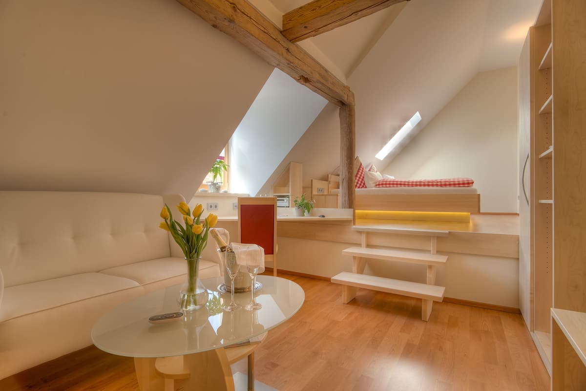 A modern loft bedroom with a sloped ceiling, a white sofa, a glass table with a vase of flowers, and a bench.