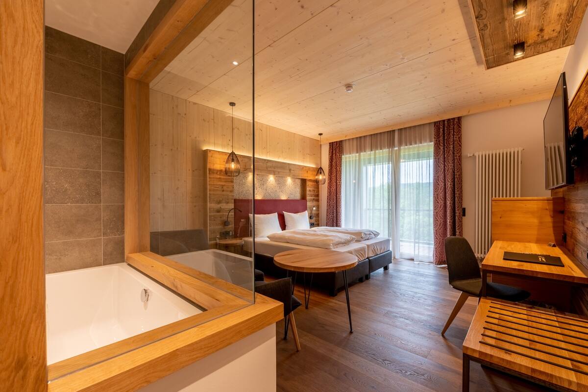 A modern hotel room with a large bed, a wooden desk, a glass-enclosed bathtub, and a view of the outdoors.