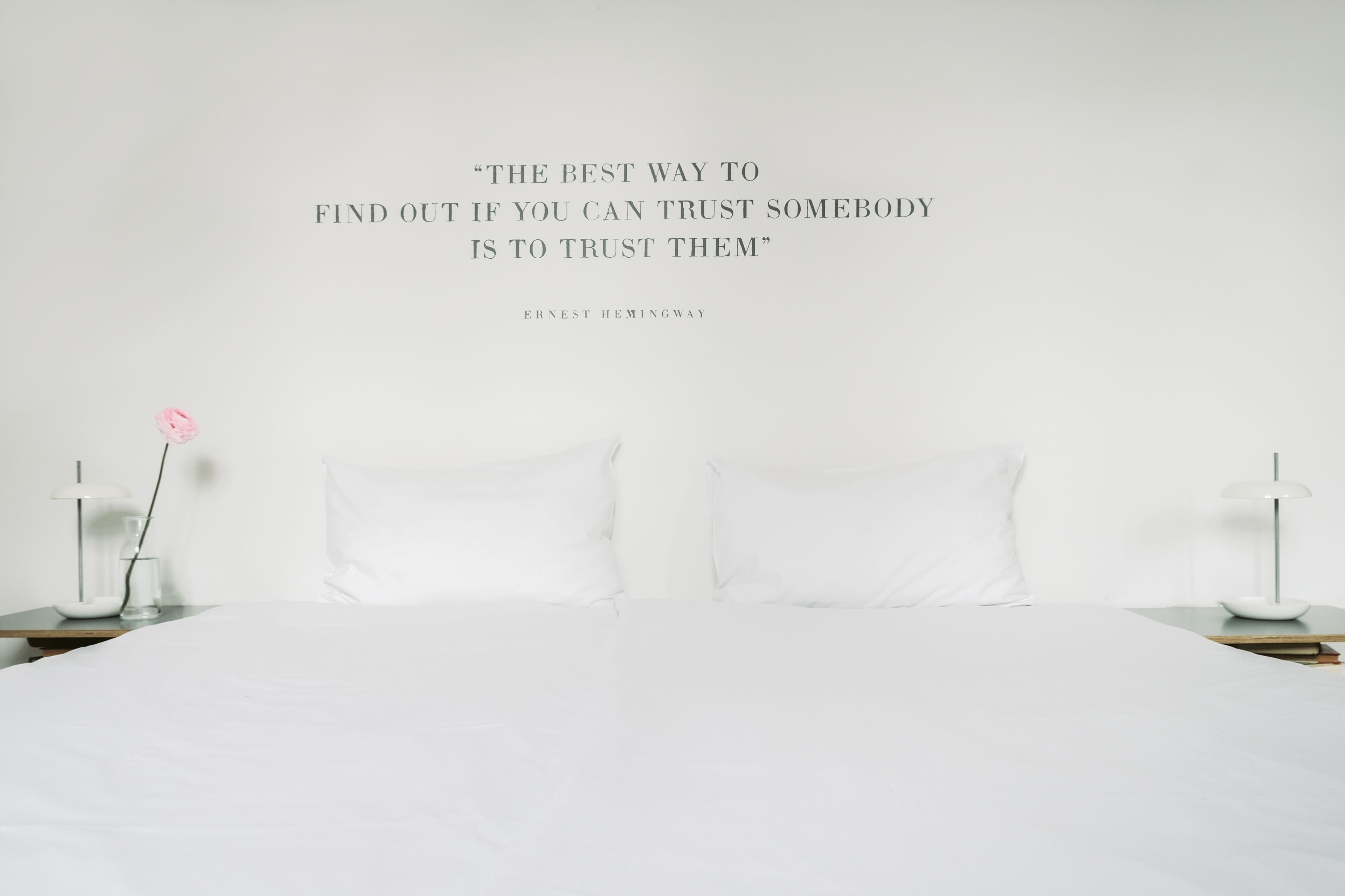 Un dormitorio minimalista con una cama grande, ropa de cama blanca y una cita de Ernest Hemingway en la pared.