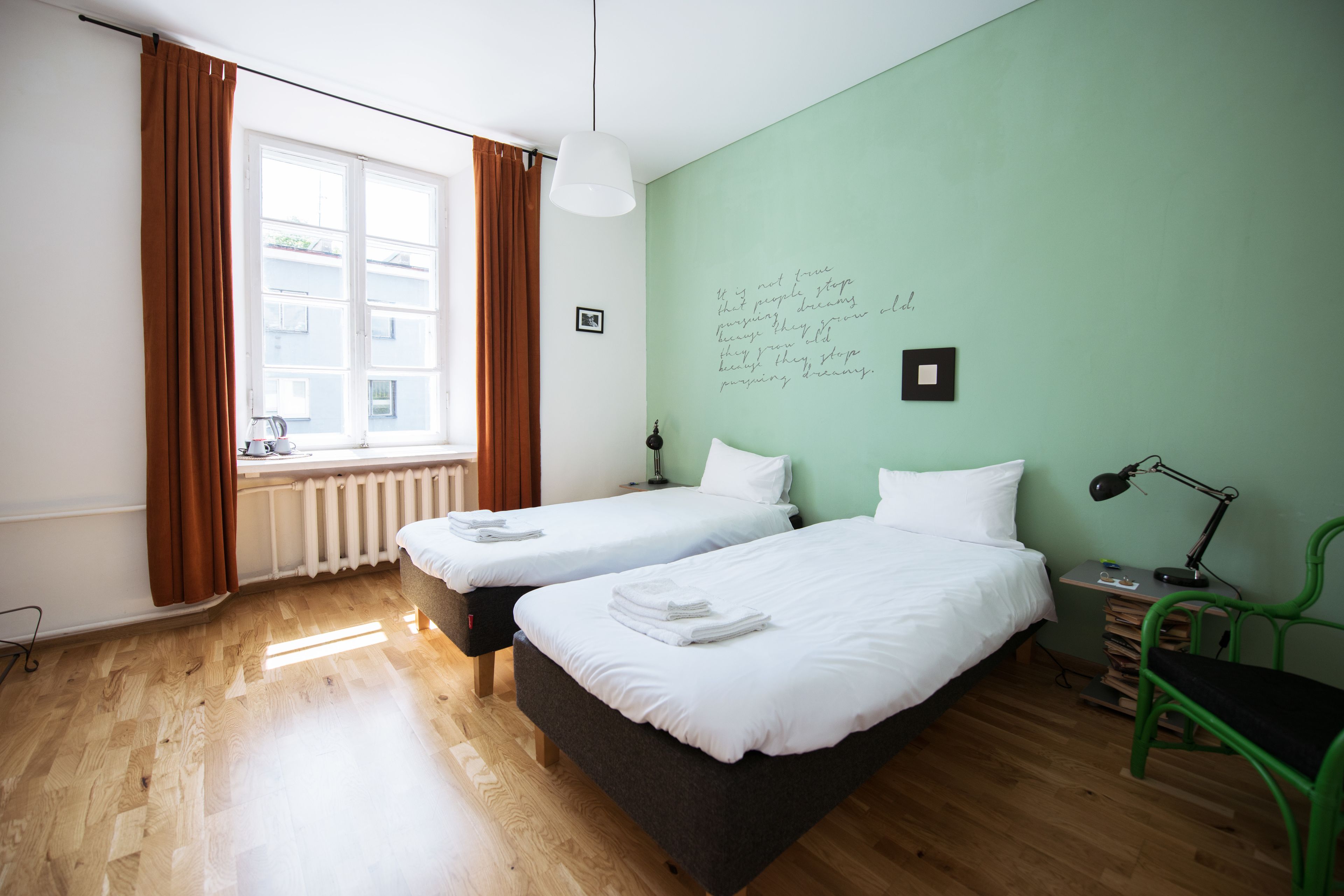 A room with two beds, a radiator, a window with curtains, a desk with a lamp, and a green chair.