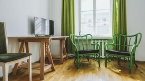 A room with a wooden table, a television, two green chairs, and a window with green curtains. - Bookinn B&B (Vilnius)