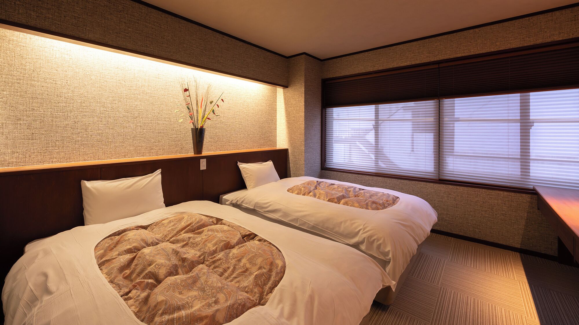 Habitación de hotel con dos camas, una mesita de noche, un florero y una ventana con persianas.