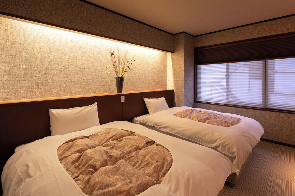 Habitación de hotel con dos camas, una mesita de noche, un florero y una ventana con persianas.