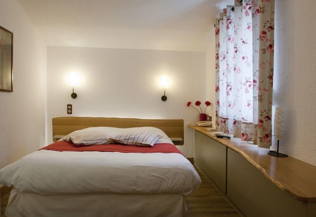 Une chambre d’hôtel avec un lit, deux lampes fixées au mur, une fenêtre avec des rideaux à motifs floraux et une table de chevet avec un téléphone et un vase de fleurs.