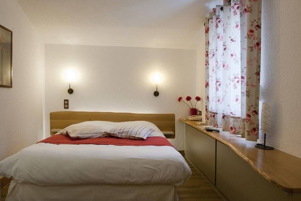 Une chambre d’hôtel avec un lit, deux lampes fixées au mur, une fenêtre avec des rideaux à motifs floraux et une table de chevet avec un téléphone et un vase de fleurs.