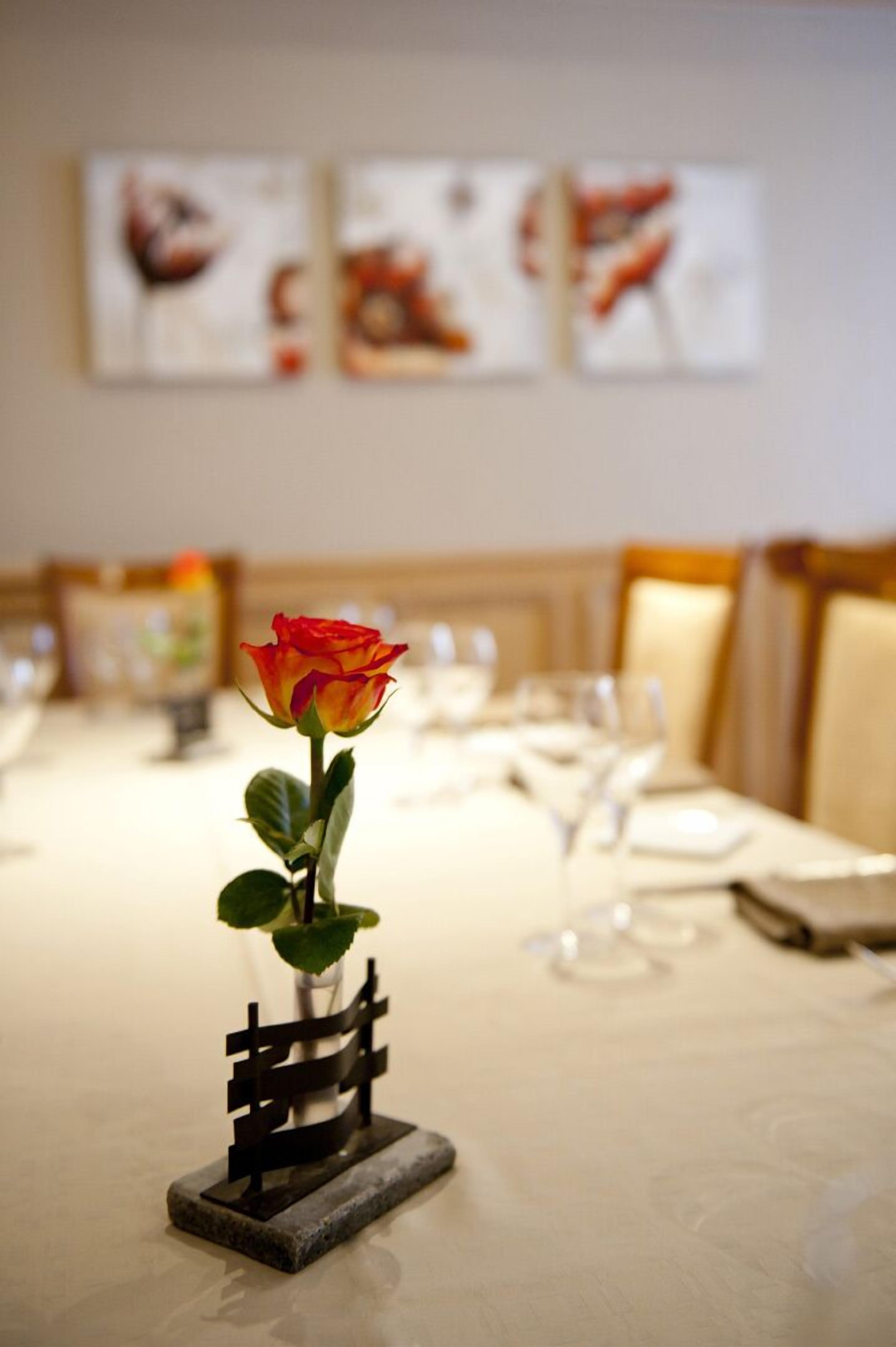 Una mesa de comedor con una rosa en un florero, un pequeño adorno similar a una cerca y un fondo desenfocado con obras de arte enmarcadas.