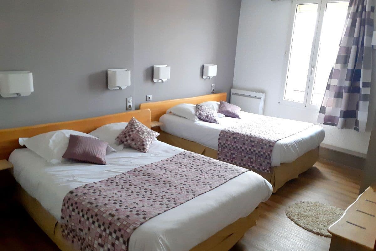 Une chambre d’hôtel avec deux lits, chacun doté d’une tête de lit en bois, de draps blancs et de plaids à motifs. On y trouve des appliques murales et une fenêtre avec un rideau.