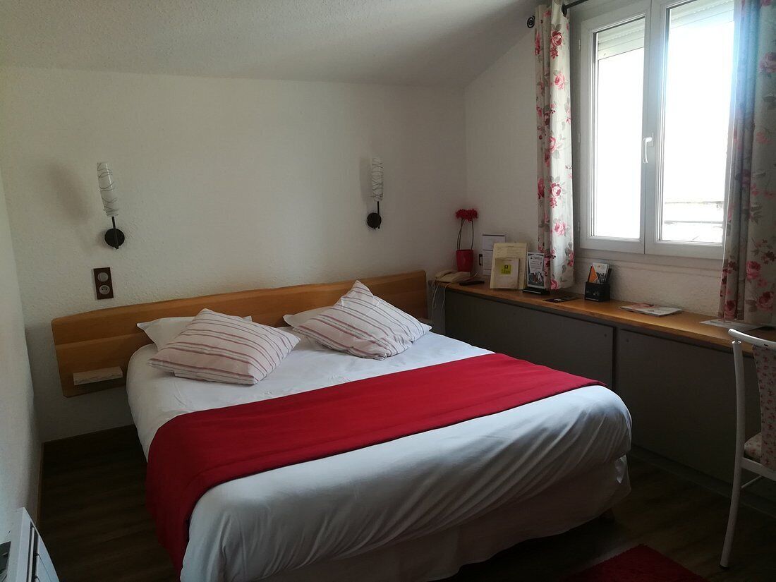 Ein Schlafzimmer mit einem Bett, einem hölzernen Kopfteil, einer roten und weißen Bettdecke, einem Fenster mit blumigen Vorhängen und einem Schreibtisch mit Stuhl.