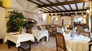 A restaurant with wooden beams, white tablecloths, and a stone fireplace. Tables are set with wine glasses and plates. - Hôtel restaurant Le Cheval Rouge (Sainte-Menehould)