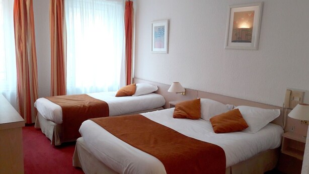 A hotel room with two beds, each with white bedding and orange throw pillows. There is a bedside table with a lamp on each side of the beds. The room has a red carpet and a window with sheer curtains. - Hôtel restaurant Le Cheval Rouge (Sainte-Menehould)
