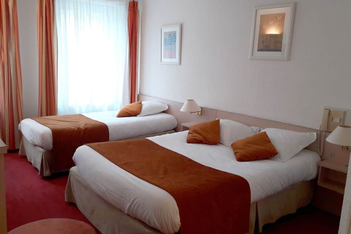 Une chambre d’hôtel avec deux lits, chacun agrémenté de draps blancs et de coussins décoratifs orange. Il y a une table de chevet avec une lampe de chaque côté des lits. La chambre est dotée d’un tapis rouge et d’une fenêtre avec des rideaux légers.