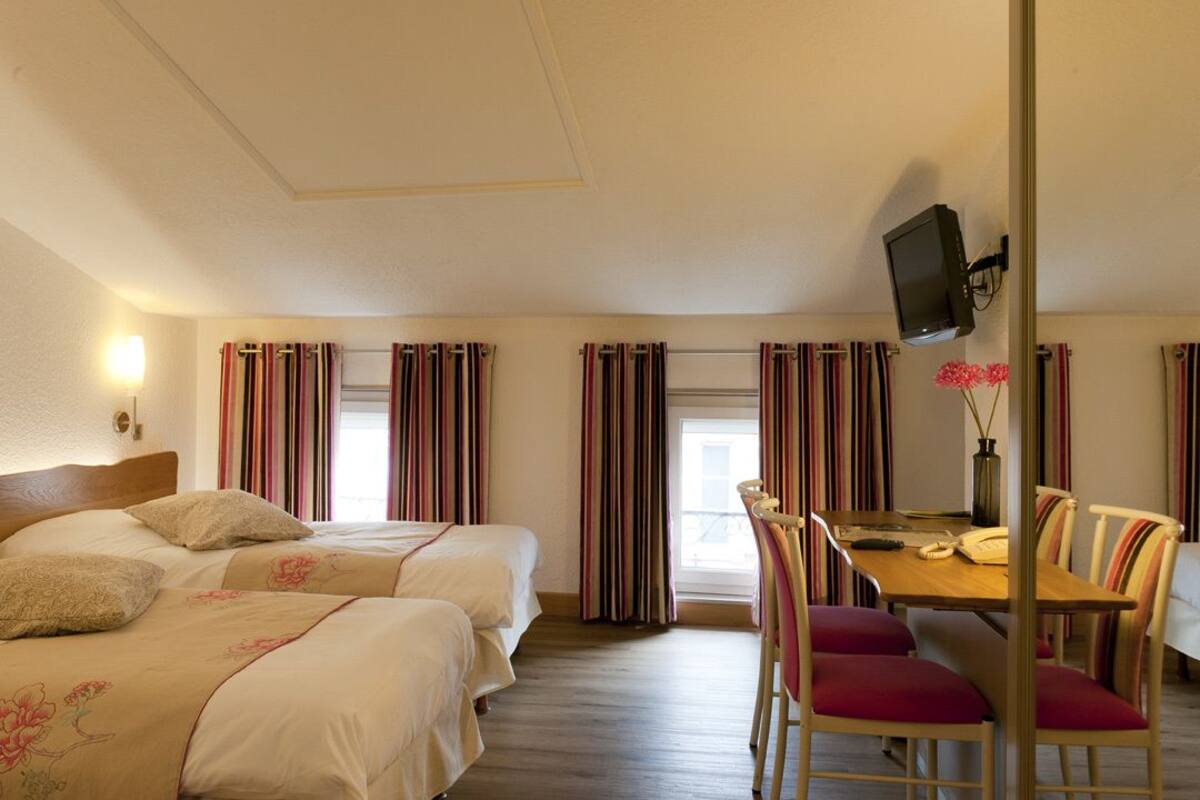Une chambre d’hôtel avec deux lits, une table à manger avec des chaises, un téléviseur fixé au mur et des rideaux à rayures.