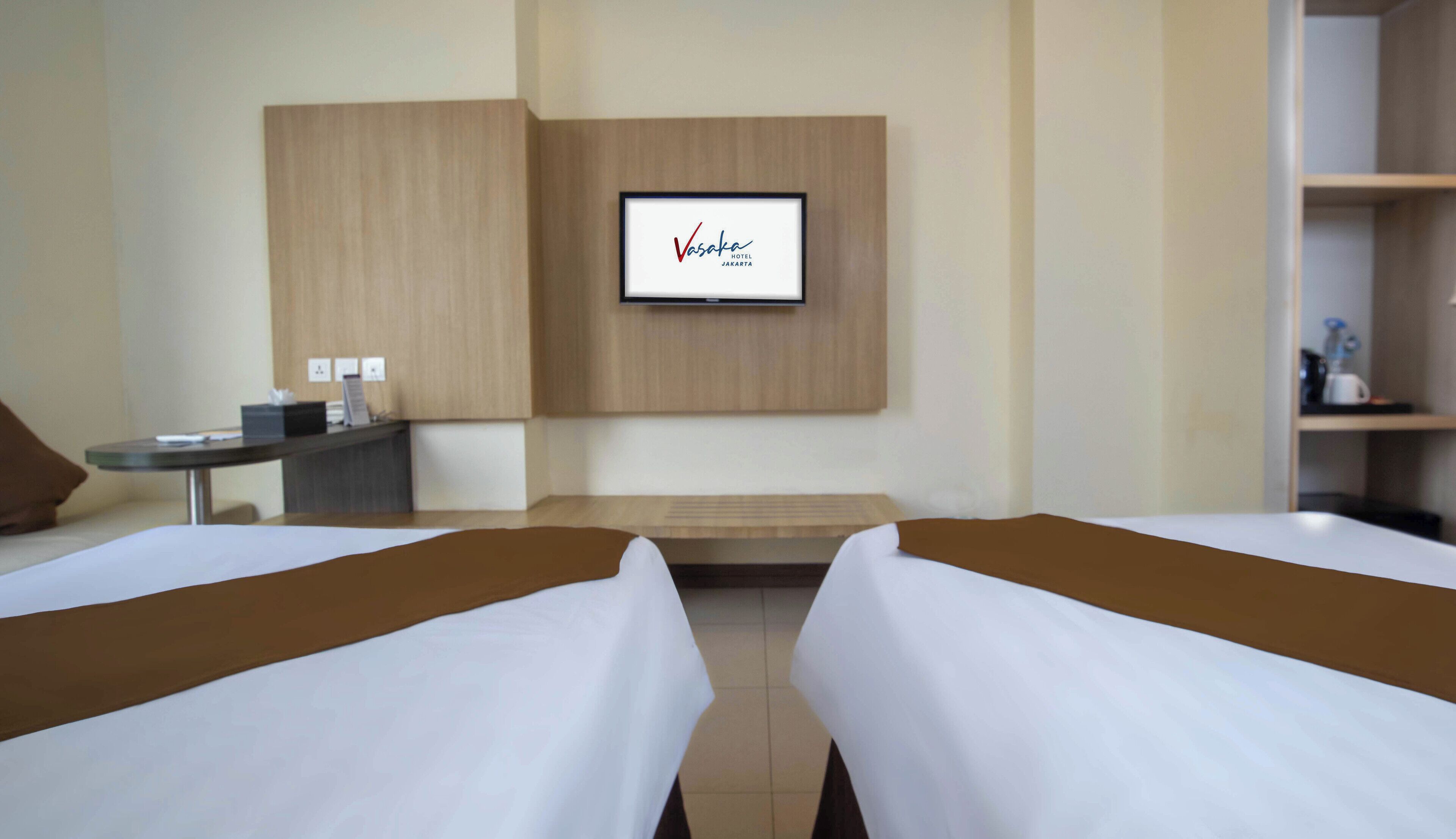 Habitación de hotel con dos camas, un escritorio, un televisor montado en la pared y una pequeña mesa con una silla.