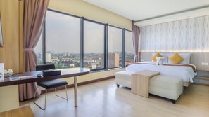Une chambre d’hôtel avec une grande fenêtre, un bureau, un lit avec une literie blanche et deux oreillers, et une petite table de chevet en bois.