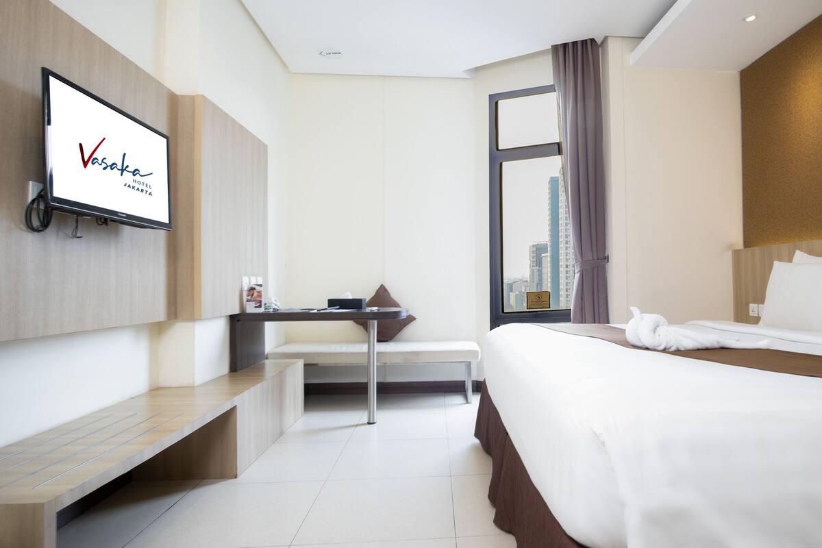 A hotel room with a bed, a desk, a chair, a TV mounted on the wall, and a window with a city view.