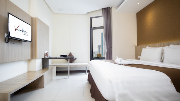 A hotel room with a bed, a desk, a chair, a TV mounted on the wall, and a window with a city view.