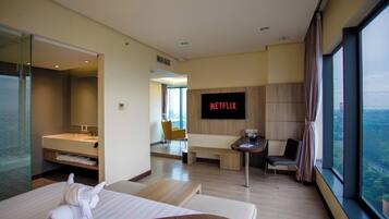 A modern hotel room with a large window, a flat-screen TV, a sofa, a small table, and a bathroom with a mirror and sink.