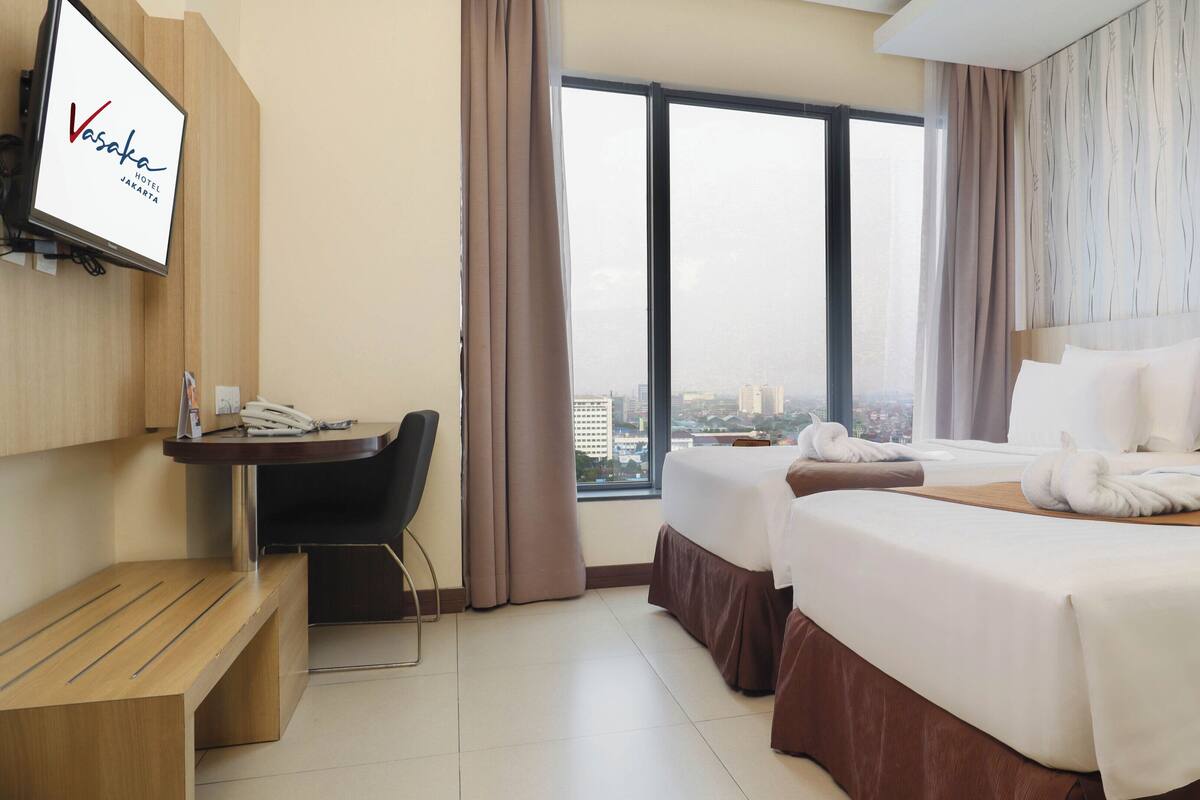 A hotel room with a desk, two beds, a city view, and a TV displaying the hotel name.