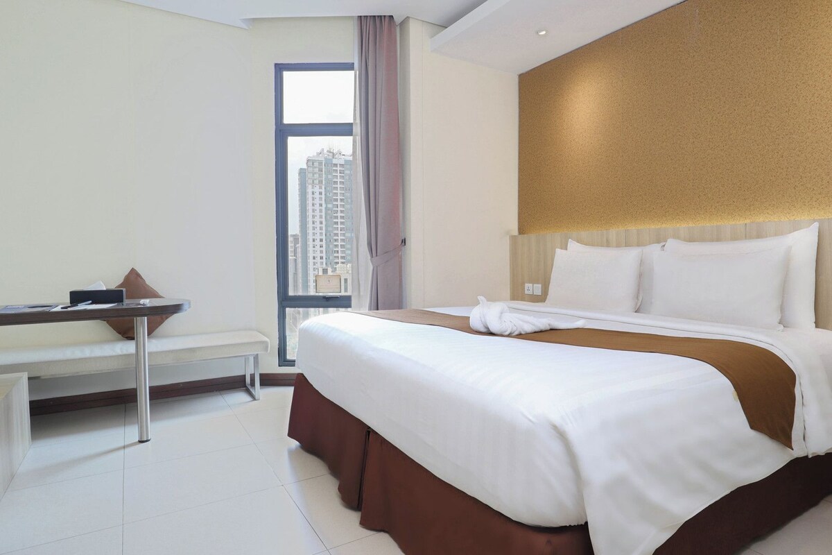 A hotel room with a bed, a desk, a chair, and a view of a cityscape through a window.