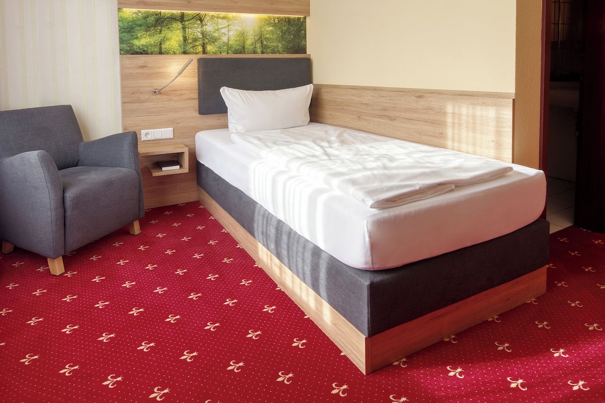 Une chambre d’hôtel bien rangée, avec un lit, un fauteuil gris, une table de chevet et un téléviseur fixé au mur.