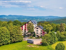 A multi-story hotel with a red and white facade, surrounded by lush greenery and a parking area.