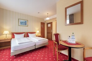 Ein Hotelzimmer mit zwei Betten, einem Schreibtisch mit Stuhl, einem runden Tisch mit roter Tischdecke und einem Spiegel an der Wand.